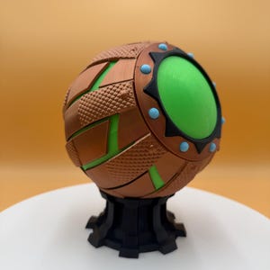 May include: A spherical, bronze-colored object with a textured surface and green accents. The sphere has a large, circular green center and is mounted on a black base. Blue and black details are also present.