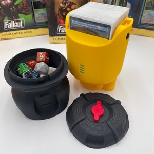 May include: A yellow card deck holder with a deck of cards inside. The holder has a black base and a red button. There is a black container with dice inside and a black lid with a red button. The card deck holder is next to two Magic: The Gathering card boxes.