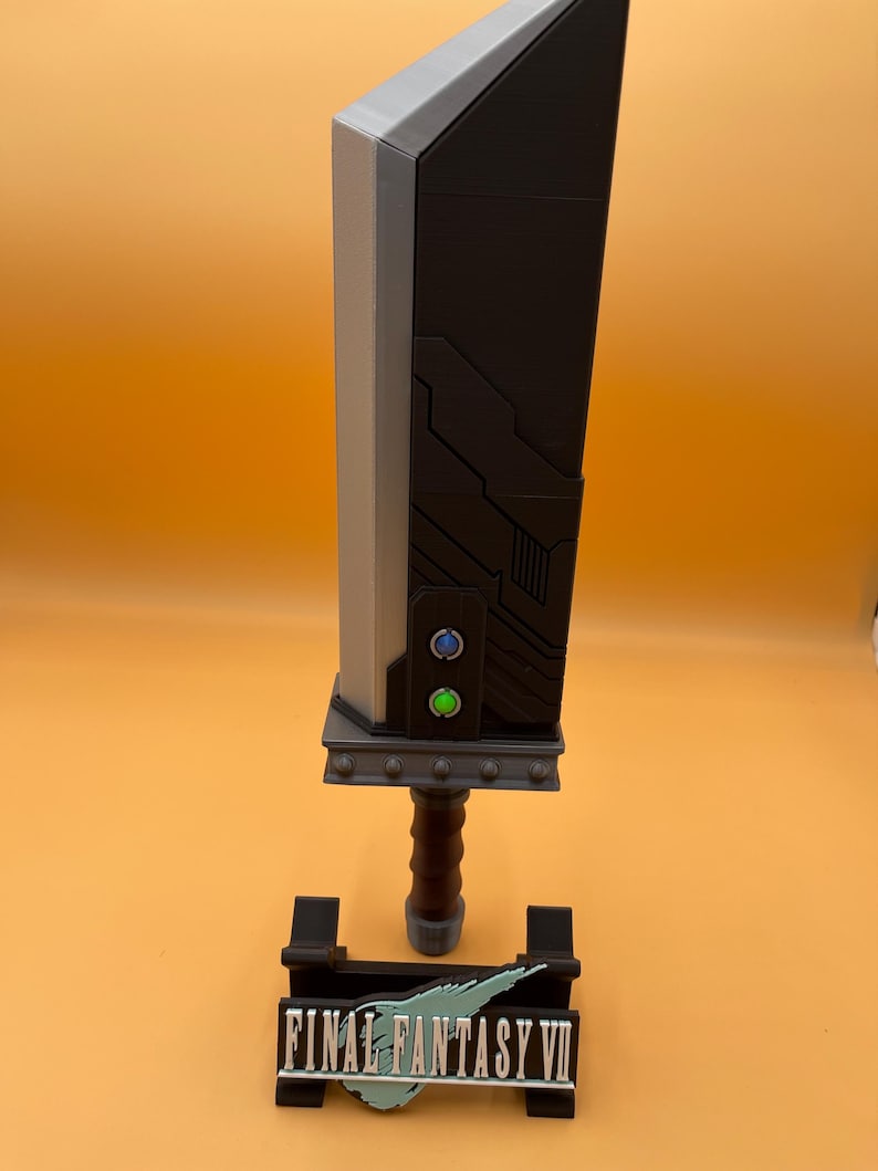 May include: A replica Buster Sword from Final Fantasy VII, featuring a large, black blade with gray accents. The hilt is brown, and the sword rests on a black stand with the game's logo. The sword has two buttons, one blue and one green.