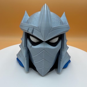Può includere: Una scultura grigio chiaro stampata in 3D del casco di Shredder dei Teenage Mutant Ninja Turtles. Il casco presenta un design dettagliato con bordi affilati, una parte superiore appuntita e una maschera nera. La base è blu.