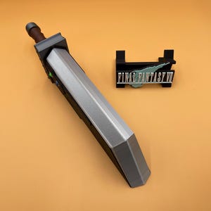 May include: A gray and silver replica of a large sword, with a brown handle and a black stand with the text "FINAL FANTASY VII". The sword is a prop from the Final Fantasy VII video game.