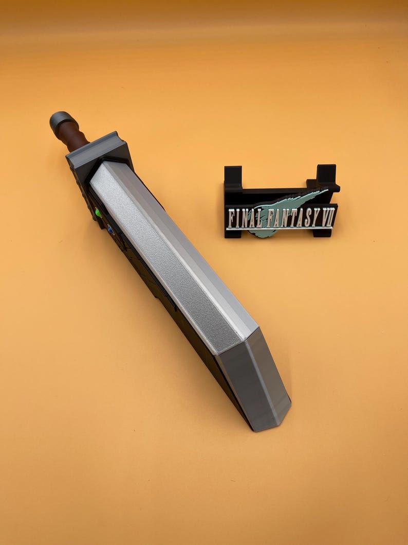 May include: A gray and silver replica of a large sword, with a brown handle and a black stand with the text "FINAL FANTASY VII". The sword is a prop from the Final Fantasy VII video game.