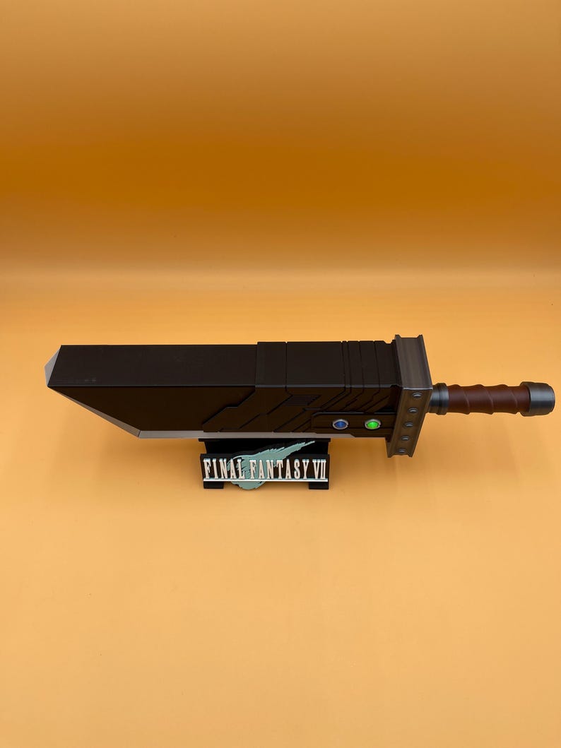 May include: A large, black Buster Sword replica from Final Fantasy VII, with a brown handle and silver accents. The sword rests on a black stand with the game's logo. The blade is angled and has a detailed design.