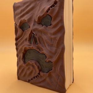 Op de afbeelding: Een bruin lederen gebonden boek met een eng gezicht gebeeldhouwd op de voorzijde. Het gezicht heeft twee ogen, een neus en een wijd open mond. Het boek is gesloten en staat rechtop op een licht oranje oppervlak.