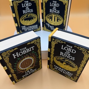 Può includere: Quattro libri in miniatura con copertine nere e scritte dorate. I libri sono intitolati "The Lord of the Rings", "The Fellowship of the Ring", "The Two Towers", "The Hobbit" e "The Return of the King". I libri hanno pagine bianche e bordi dorati.