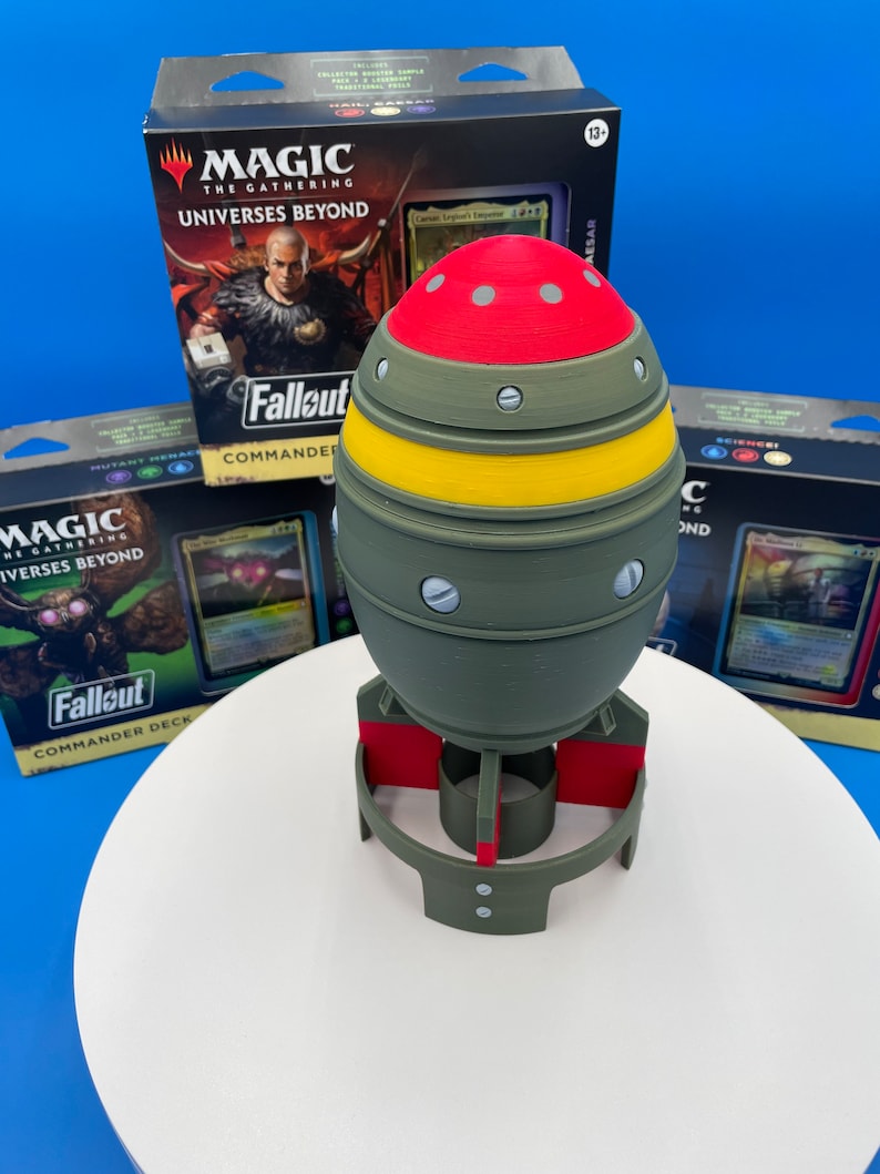 May include: A 3D printed model of a green and red bomb with a yellow stripe. The bomb is sitting on a white circular base. The bomb is surrounded by three Magic: The Gathering card boxes. The boxes are for the "Universes Beyond" set and feature the Fallout theme.