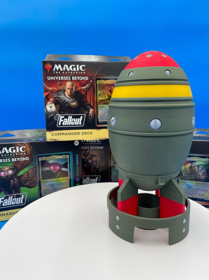 May include: A green, yellow, and red 3D printed model of a bomb. The bomb is sitting on a white surface. The bomb is surrounded by three Magic: The Gathering card decks. The card decks are all from the "Universes Beyond" set and feature the Fallout logo.