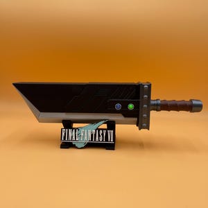 May include: A large, black and silver Buster Sword replica from Final Fantasy VII, with a brown handle and green and blue accents. The sword rests on a black stand with the game's logo.