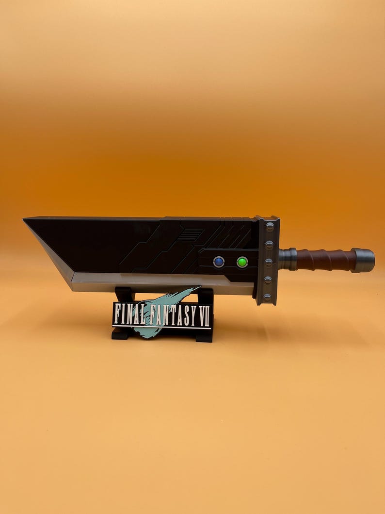 May include: A large, black and silver Buster Sword replica from Final Fantasy VII, with a brown handle and green and blue accents. The sword rests on a black stand with the game's logo.