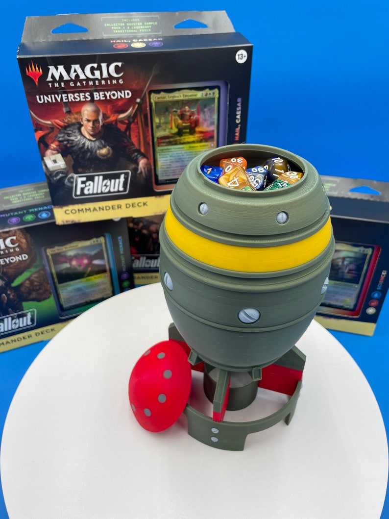 May include: A green and yellow bomb-shaped dice tower with a red lid and dice inside. The dice tower is sitting on a white surface. The tower is a 3D printed object. The tower is surrounded by three Magic: The Gathering card boxes. The boxes are for the Fallout Commander Deck.