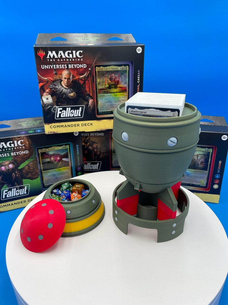 May include: A green and red bomb-shaped dice tower with a removable lid. The tower is filled with colorful dice. The tower is sitting on a white surface next to a box of Magic: The Gathering cards. The box has the text "Universes Beyond" and "Fallout" on it.