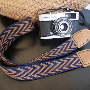 May include: A navy blue and tan chevron patterned camera strap with leather ends. An Olympus Trip 35 camera sits behind the strap. A woven straw bag is in the background.