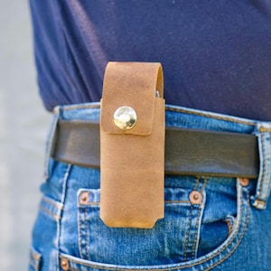 Może przedstawiać: Brązowe skórzane etui ze srebrnym zapięciem na zatrzask, przymocowane do czarnego paska. Etui jest noszone w talii, na niebieskich dżinsach. Skóra ma teksturowany wygląd.