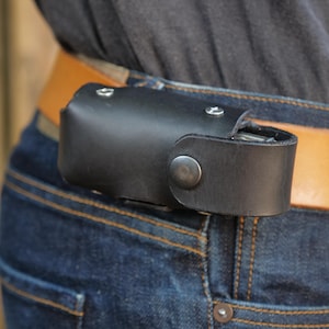 May include: A black leather belt holster featuring a secure snap button closure and silver-toned metal rivets. The rectangular pouch is designed to be worn on a tan leather belt, which is threaded through the loops of dark blue denim jeans.