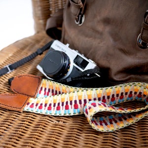 May include: A colourful camera strap with a brown leather strap and a silver camera. The strap has a geometric pattern with red, yellow, blue and green colours.