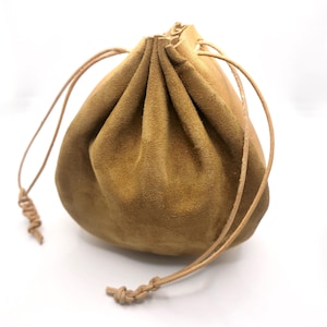 Könnte beinhalten: Ein beigefarbener Beutel aus Wildleder mit Kordelzug und einer weichen, matten Textur. Die Tasche verfügt über einen gerafften Verschluss, der durch dünne, helle Lederbänder gesichert ist, die seitlich herausragen und in einfachen Knoten enden.