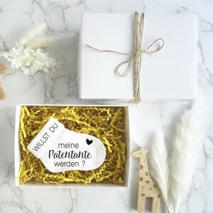 May include: White gift box with a small white sock with the text "Willst du meine Patenante werden?" and a black heart. The box is filled with yellow crinkle paper.