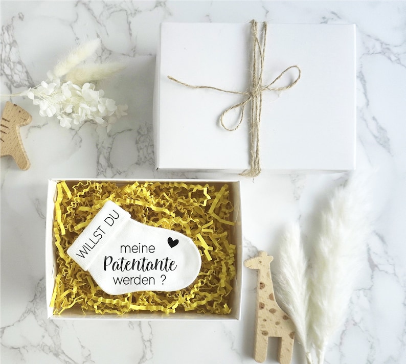 May include: White gift box with a small white sock with the text "Willst du meine Patenante werden?" and a black heart. The box is filled with yellow crinkle paper.