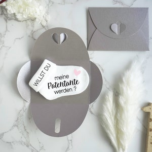 May include: A white baby sock with the text "Willst du meine Patenante werden?" and a pink heart. The sock is inside a gray envelope with a heart cut-out.