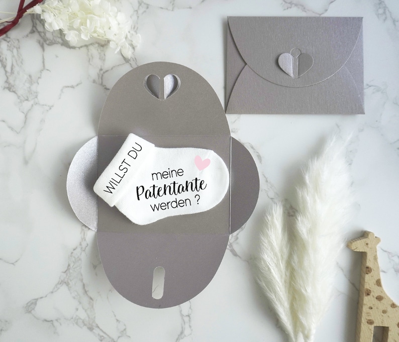 May include: A white baby sock with the text "Willst du meine Patenante werden?" and a pink heart. The sock is inside a gray envelope with a heart cut-out.