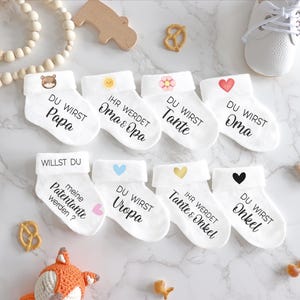 May include: Set of 8 white baby socks with different designs and text. The socks say "Du wirst Papa", "Ihr werdet Oma & Opa", "Du wirst Tante", "Du wirst Oma", "Willst du meine Patentante werden?", "Du wirst Uropa", "Ihr werdet Tante & Onkel", and "Du wirst Onkel".