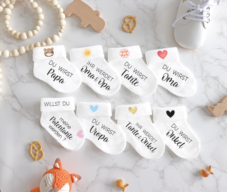 May include: Set of 8 white baby socks with different designs and text. The socks say "Du wirst Papa", "Ihr werdet Oma & Opa", "Du wirst Tante", "Du wirst Oma", "Willst du meine Patentante werden?", "Du wirst Uropa", "Ihr werdet Tante & Onkel", and "Du wirst Onkel".