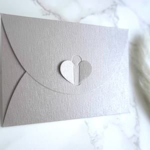 May include: A light gray envelope with a heart-shaped cutout. The cutout is filled with two overlapping hearts, one white and one gray.