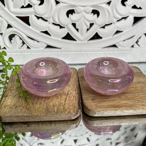 Peut inclure: Deux bougeoirs en verre rose iridescent avec une forme arrondie en forme de bol. Les bougeoirs sont posés sur deux sous-verres carrés en bois.