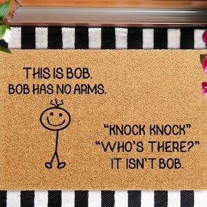 May include: A rectangular doormat with the text "THIS IS BOB. BOB HAS NO ARMS." and a stick figure. Below, the mat reads "KNOCK KNOCK" "WHO'S THERE?" "IT ISN'T BOB." The mat is brown with a black and white striped border.