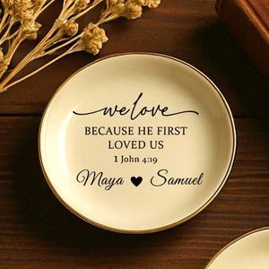 May include: Two small, off-white ring dishes with gold rims, inscribed with the words "we love" and a Bible verse. The names Maya and Samuel are on one dish, and Scarlett and Andrew on the other. Gold rings and a candlestick are also visible.
