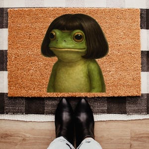 May include: A doormat featuring a green frog with a dark brown bob haircut. The frog is centered on a rectangular, light brown mat, set against a black and white checkered background. Black boots are visible at the bottom.