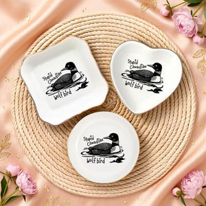 May include: Three white ceramic dishes with a gold trim, each featuring a black and white illustration of a loon and the text "Stupid Canadian Wolf bird". The dishes are in square, heart, and round shapes.