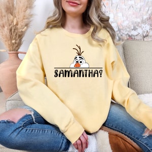 Puede incluir: Una sudadera amarilla con una imagen de dibujos animados de Olaf de Frozen asom&aacute;ndose por encima de una l&iacute;nea con el texto "SAMANTHA?" impreso debajo.