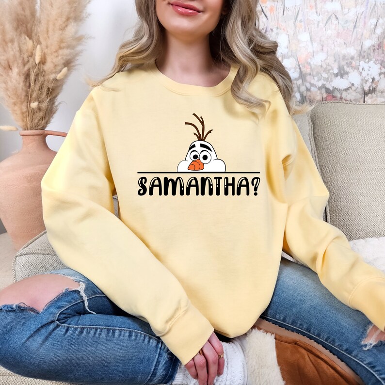 Puede incluir: Una sudadera amarilla con una imagen de dibujos animados de Olaf de Frozen asom&aacute;ndose por encima de una l&iacute;nea con el texto "SAMANTHA?" impreso debajo.