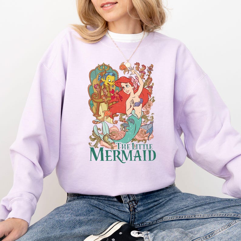 Puede incluir: Una sudadera lila con un gr&aacute;fico colorido de Ariel y Flounder de La Sirenita. El gr&aacute;fico incluye el texto "THE LITTLE MERMAID" en verde. La sudadera es de cuello redondo.