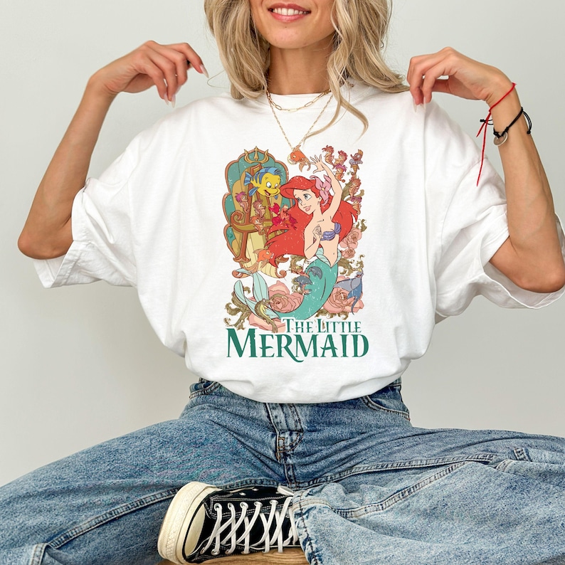Puede incluir: Camiseta blanca con un gr&aacute;fico colorido de Ariel de La Sirenita. El dise&ntilde;o incluye a Ariel, Flounder y elementos florales, con la frase "THE LITTLE MERMAID" en verde. Una camiseta de estilo casual y c&oacute;modo.