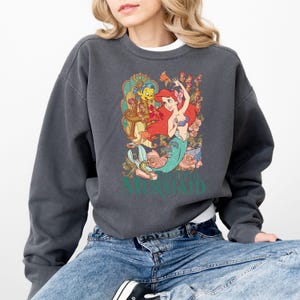Puede incluir: Sudadera gris oscuro con un dise&ntilde;o de Ariel de La Sirenita, con Flounder y detalles florales. El dise&ntilde;o incluye el texto "The Little Mermaid" en verde. La sudadera tiene mangas largas y cuello redondo.