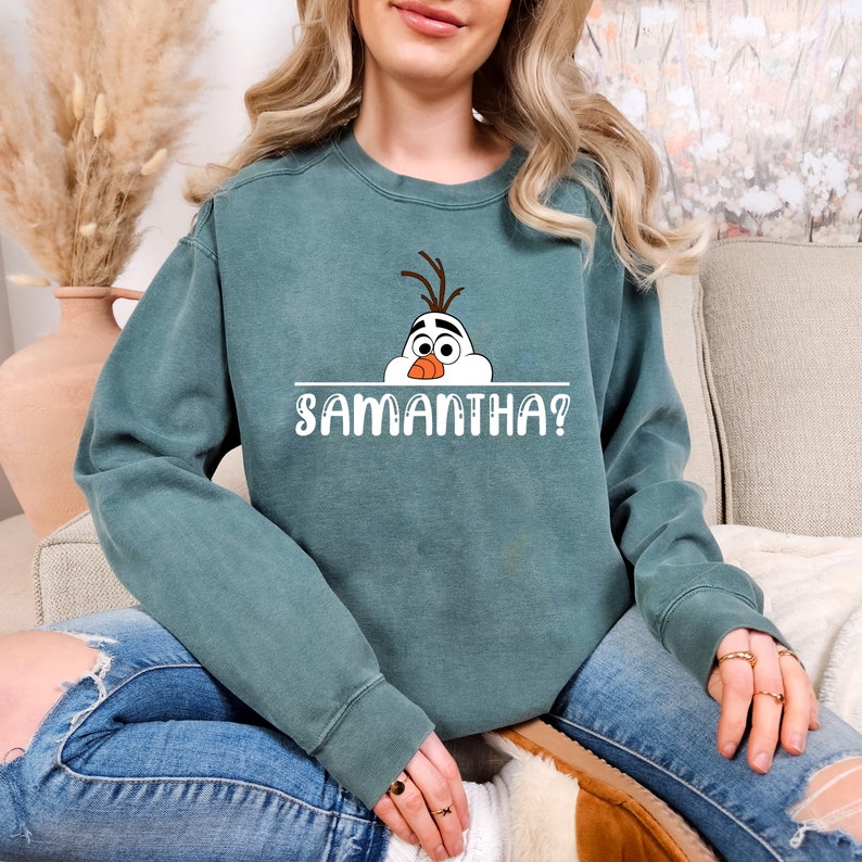 Puede incluir: Una sudadera azul verdosa con una imagen de dibujos animados de Olaf de Frozen asom&aacute;ndose por encima de una l&iacute;nea que dice "SAMANTHA?"