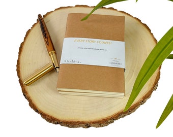 May include: A brown notebook with the text "EVERY STORY COUNTS!" and "Thank you for traveling with us" on the cover. The notebook is sitting on a wooden surface with a wooden pen next to it.