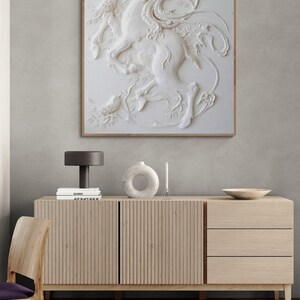 Puede incluir: Una escultura de pared tridimensional blanca de un ciervo con un p&aacute;jaro posado en sus astas. La escultura est&aacute; enmarcada y colgada en una pared gris claro sobre un gabinete de madera.