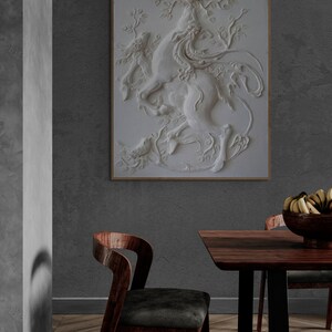 Puede incluir: Una escultura en relieve de yeso blanco de una criatura m&iacute;tica con un p&aacute;jaro posado en su cabeza, colocada contra una pared gris en un comedor moderno con una mesa y sillas de madera.