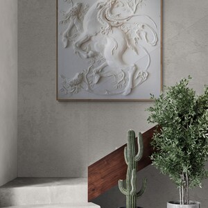 Puede incluir: Una escultura en relieve de yeso blanco de una mujer montando un caballo, enmarcada en un marco de madera clara, colgada en una pared gris. La escultura est&aacute; rodeada de plantas en macetas.