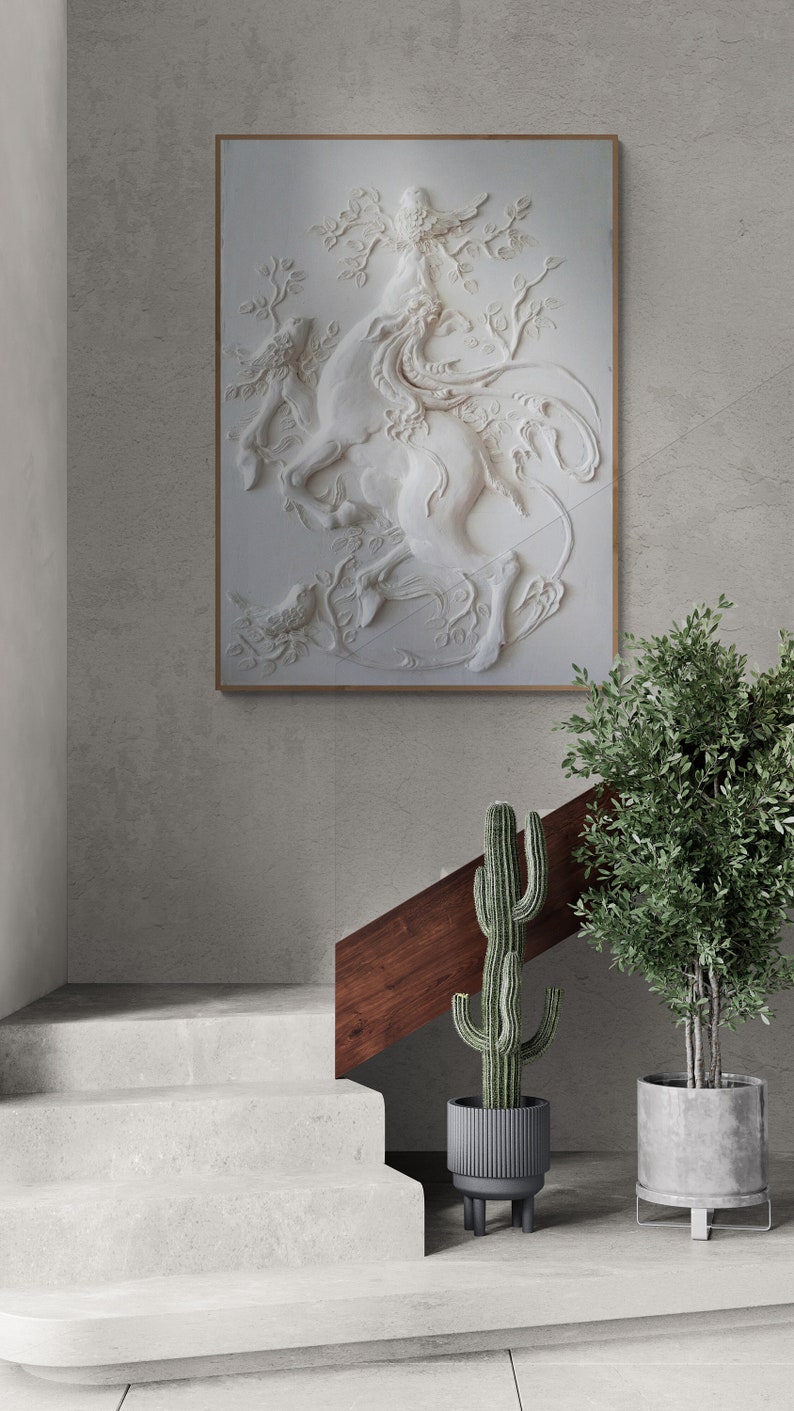 Puede incluir: Una escultura en relieve de yeso blanco de una mujer montando un caballo, enmarcada en un marco de madera clara, colgada en una pared gris. La escultura est&aacute; rodeada de plantas en macetas.