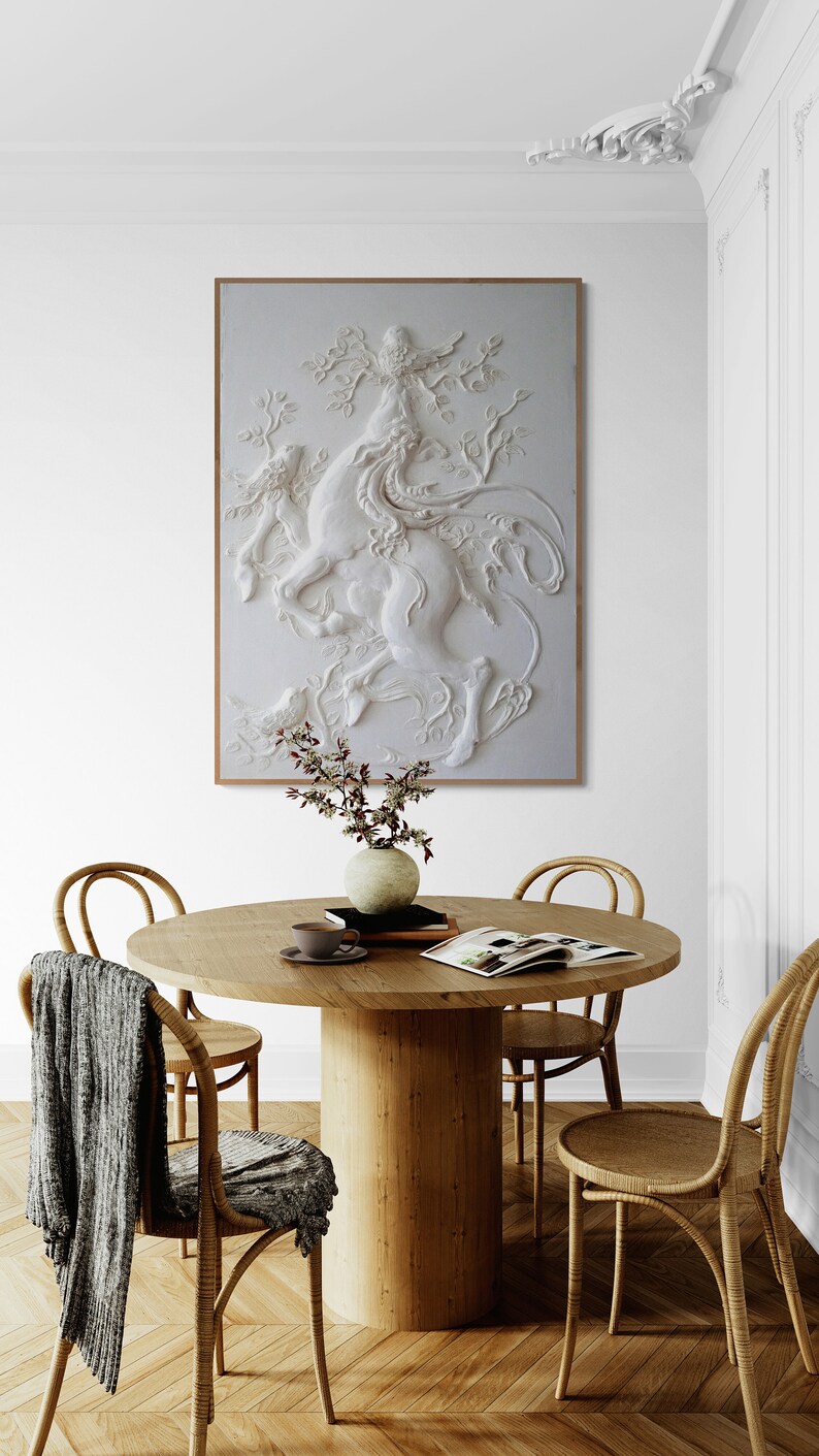 Puede incluir: Una escultura blanca en tres dimensiones de una criatura m&iacute;tica con alas, sobre un fondo blanco, cuelga sobre una mesa de madera con cuatro sillas. La mesa est&aacute; decorada con un jarr&oacute;n de flores, una taza y un platillo, y un libro.