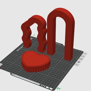 May include: A red 3D-printed heart-shaped object with a scalloped edge, accompanied by two red arch-shaped structures. The objects are on a black grid surface, likely a 3D printer bed. The design is simple and geometric.