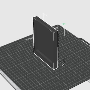 Op de afbeelding: Een donkergrijs rechthoekig object met een witte omtrek, rechtopstaand op een oppervlak met een rasterpatroon. Het object heeft een verzonken gebied langs één rand. De achtergrond is lichtgrijs.