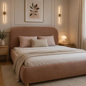 May include: A rounded, upholstered bed frame in a muted terracotta fabric with a white mattress, two light pink square pillows, and a textured beige lumbar pillow. A light beige throw blanket is draped over the foot of the bed.