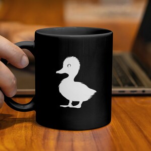 Könnte beinhalten: Schwarze Keramik-Tasse mit einer weißen Enten-Silhouette. Die Ente ist ein einfaches Cartoon-Design. Die Tasse wird von einer Hand gehalten, mit einem Laptop im Hintergrund. Die Tasse ist für Getränke geeignet.
