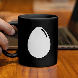 Könnte beinhalten: Schwarze Keramik-Tasse mit einem weißen Eier-Design. Die Eier-Grafik ist eine einfache, saubere Illustration. Die Tasse hat einen Standardgriff und steht auf einer Holzoberfläche mit einem Laptop im Hintergrund.