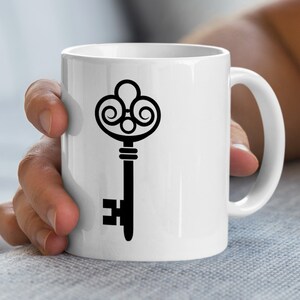 Könnte beinhalten: Weiße Keramik-Tasse mit einem schwarzen Schlüssel-Design. Der Schlüssel hat eine verzierte Oberseite mit Wirbeln und eine einfache Schlüssellochform. Die Tasse hat einen gebogenen Griff und wird von einer Person gehalten.